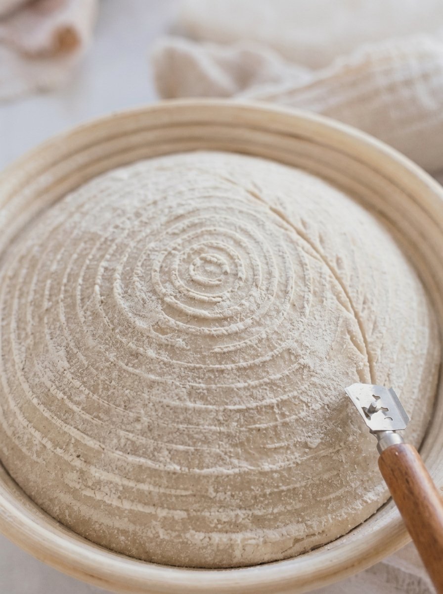 Shaped sourdough dough proofing in a banneton with a scoring tool beside it