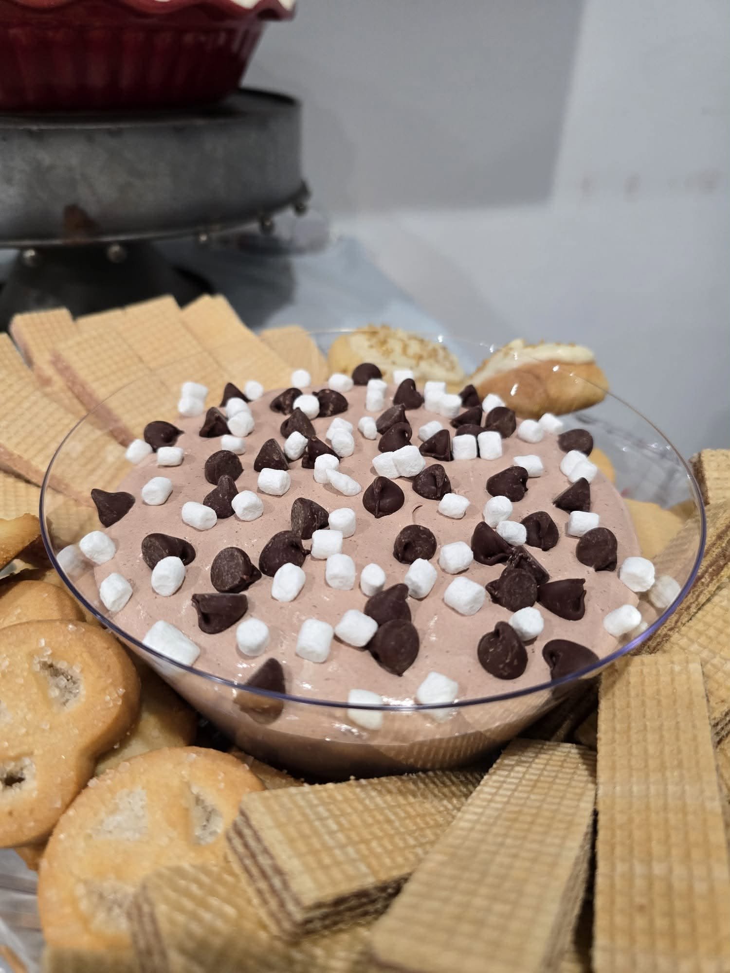 A chocolate dessert dip topped with marshmallows and chocolate chips, surrounded by cookies and wafers