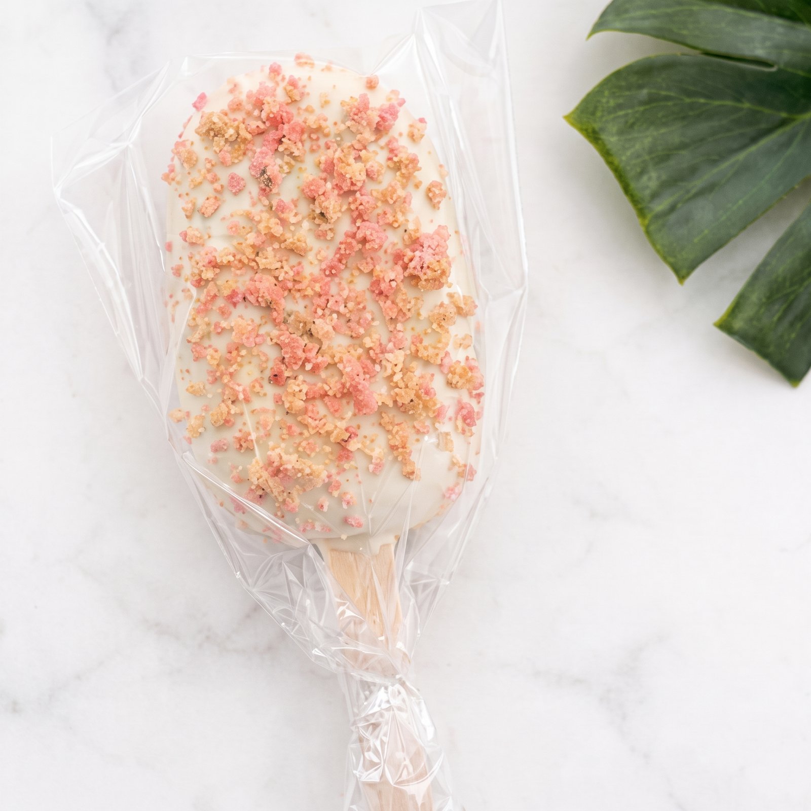 A wrapped white chocolate cakesicle topped with strawberry crunch